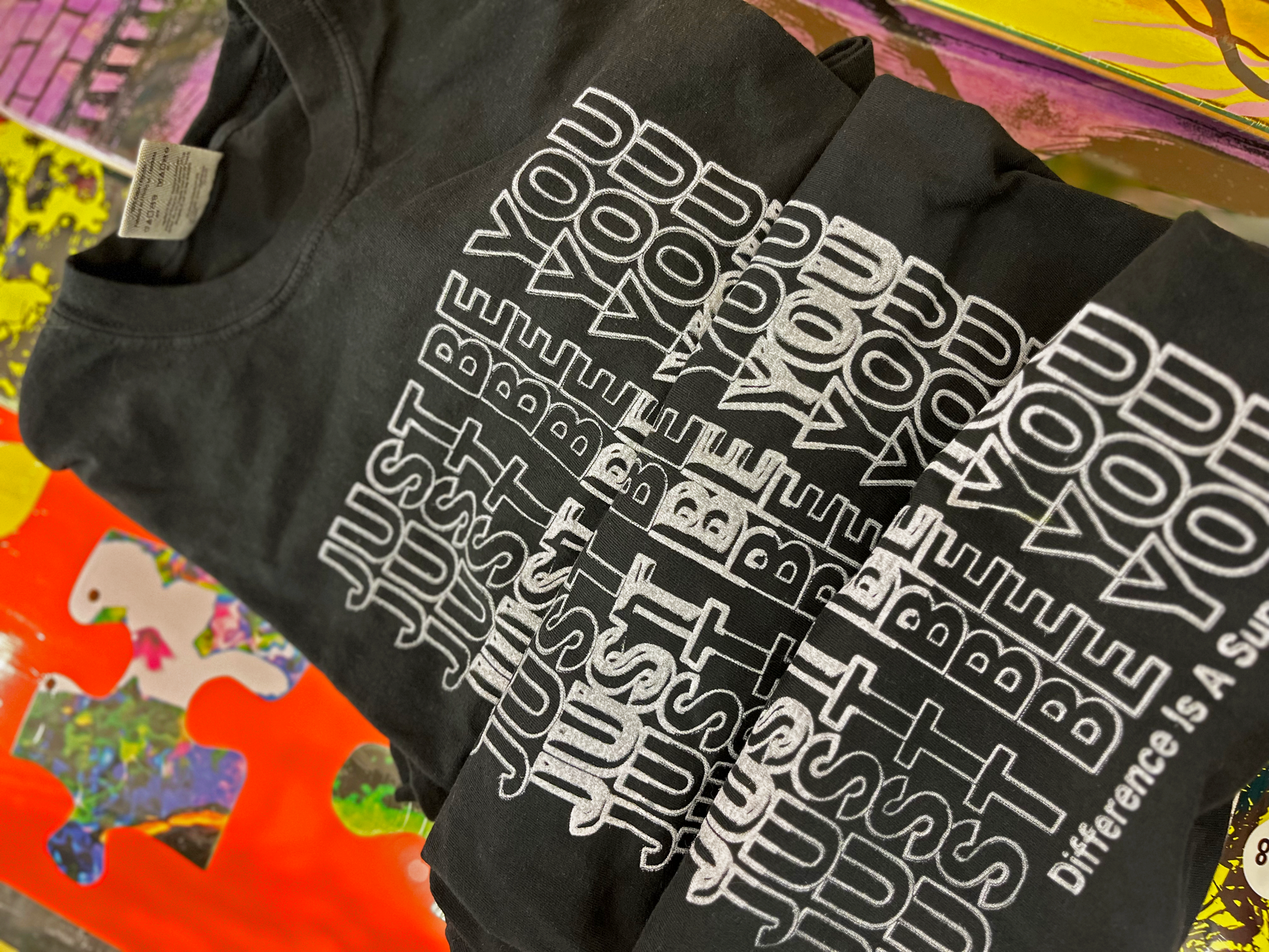 Stack of black t-shirts featuring a design that says "Just Be You" repeated seven times, and "Difference is a Superpower" in smaller text underneath. The design is white. 